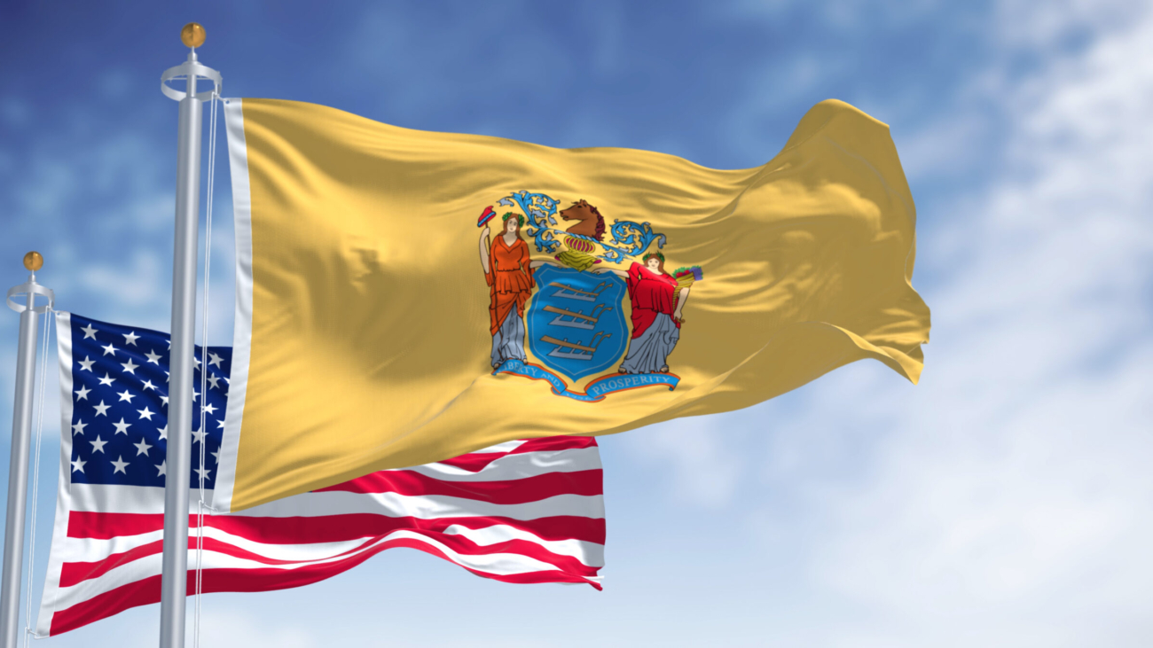 NJ State Flag with US Flag behind it flowing in the wind. NJ Flag is yellow, blue and red.