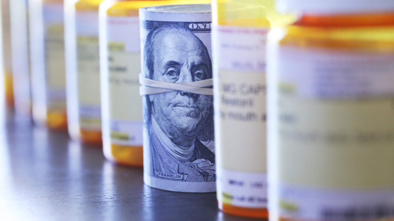 A roll of one hundred dollar bills sits among a row of prescription medication bottles. Photographed with a very shallow depth of field with the focus on the roll of money.