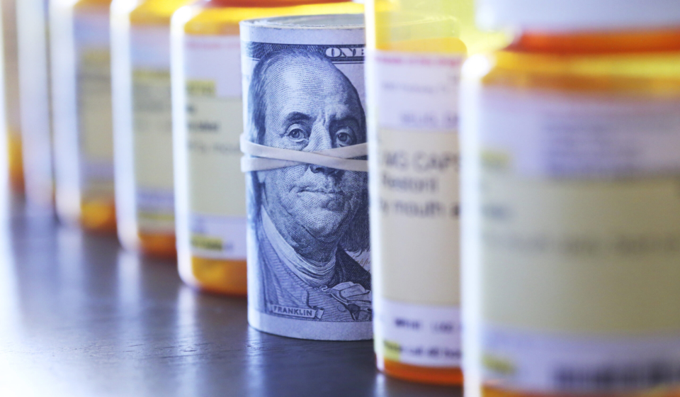 A roll of one hundred dollar bills sits among a row of prescription medication bottles. Photographed with a very shallow depth of field with the focus on the roll of money.